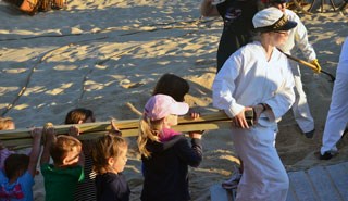 A young woman dressed as a surfman in white  carries a ladder with the help of many children.