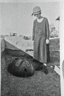 black and white image of woman in 1920s clothing looking at a large olla pottery artifact