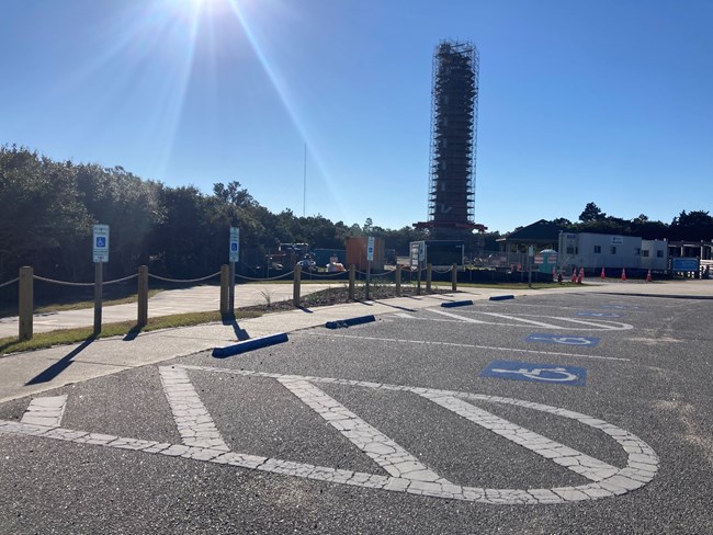 Accessible-marking parking at Cape Hatteras Lighthouse. In the foreground are four spots painted and signed with the universal symbol of accessibility. In the background, Cape Hatteras Lighthouse is surrounded yb scaffolding while undergoing restoration.