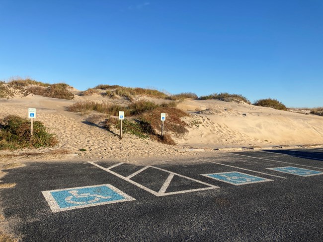 Accessible marked parking at ORV Ramp 2. Three spots painted with the universal symbol of accessibility and one access aisle connect to a sand dune overflowing into the parking spaces.