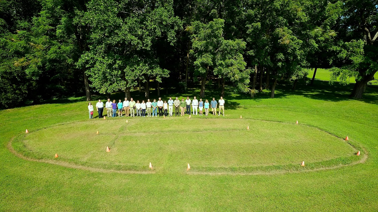 A group of people pose for a photo in a grassy lawn where a large oval is marked with cones.