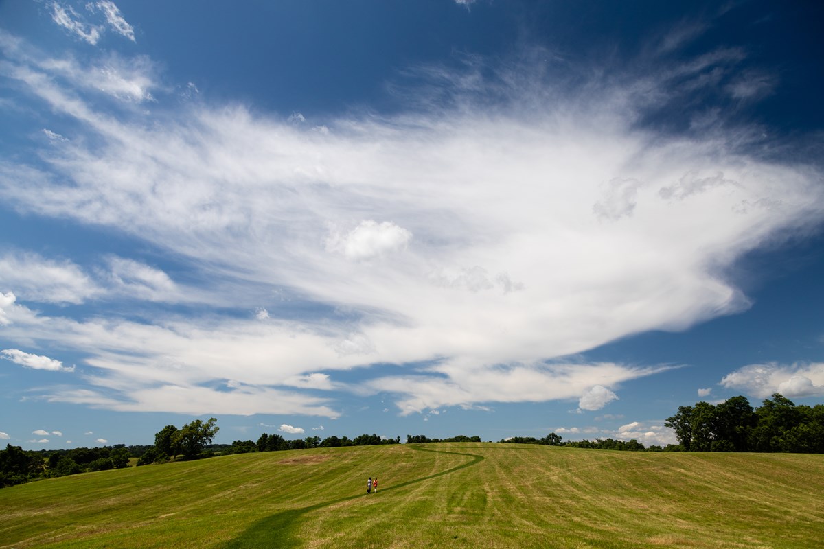 A rolling, grassy field with two hikers below a blue, cloudy sky.
