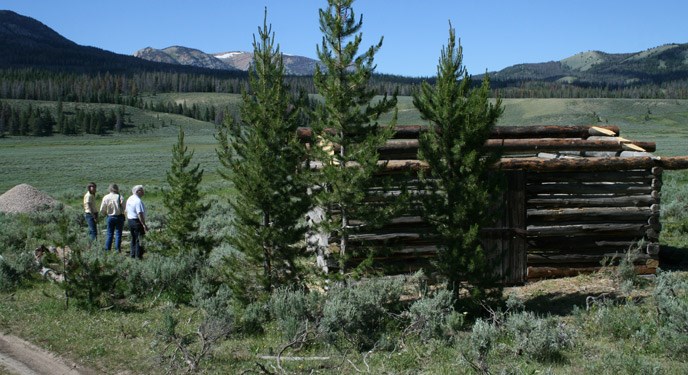 Three men stand in front of a log cabin at Fort Piney in Wyoming with mountains in background and pine trees.