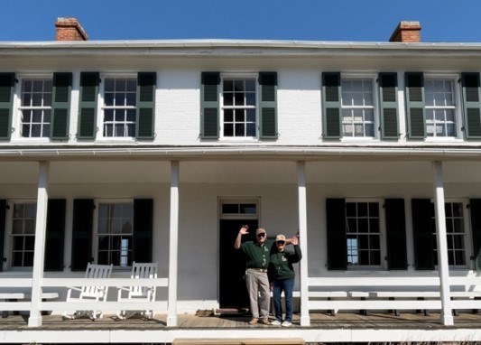 Two people stand on a porch, waving.