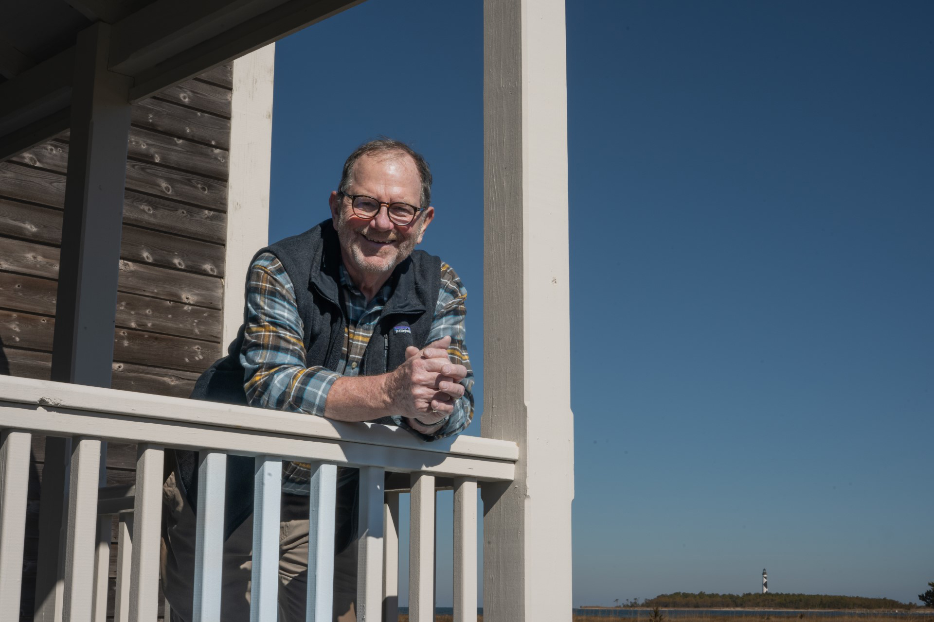 Dr. Stan Rule leans over the balancy of a building, with the Cape Lookout Lighthouse in the bottom right.