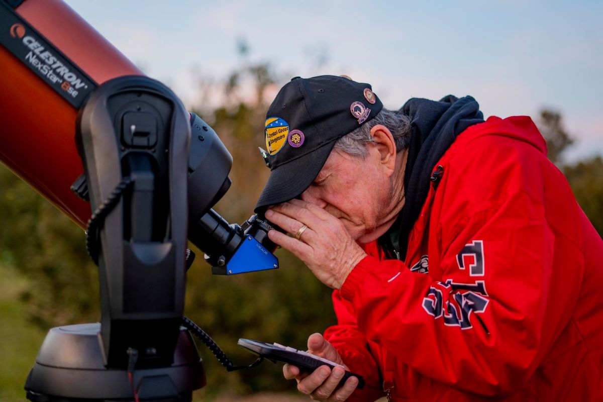A crystal coast stargazer looks through a celestron telescope. He uses his left hand to adjust the scope, and his right hand holds the remote.