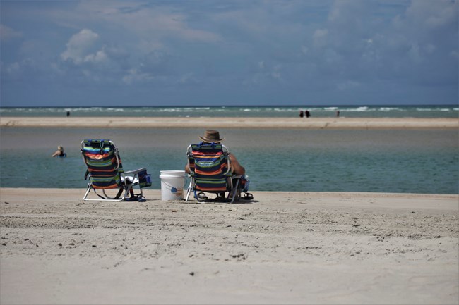 A person sits in a chair, next to another chair. Water and sand in the background.
