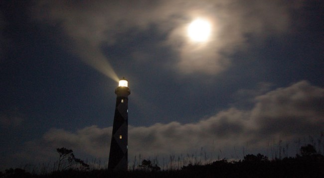 Cape Lookout Lighthouse at night with a moon in the sky and light showing from the lantern room at the top