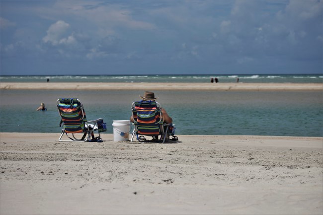 Two stripped chairs with a 5 gallon bucket between them on the sand. A person wearing a hat sits in the right chair. People in the background standing in the water. A cloudy blue sky in the background,