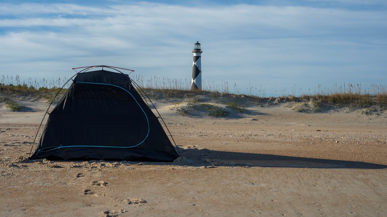 Black tent with blue lines on the left, Cape Lookout Lighthouse in the background. Grass covered dunes in between the tent and lighthouse. Blue sky with clouds in the background.