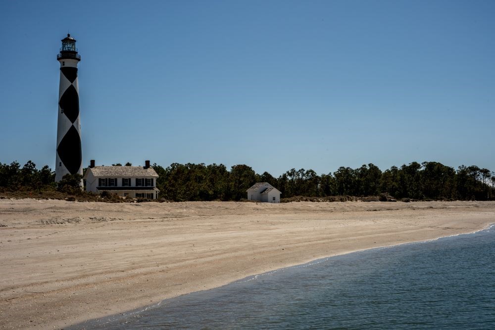 Cape Lookout Lighthouse, Keeper's Quarters, and summer kitchen with tall pine trees behind them. A blue sky is in the background. Water and sand is in the foreground.