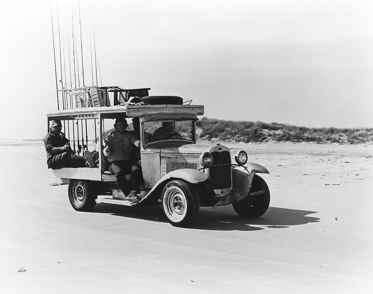 A black and white photo of 3 people sitting in a beach vehicle on the sand.