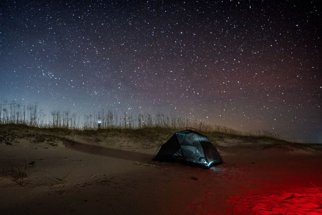 A red light shines on a tent, with dunes in the background. The night sky is covered in stars.