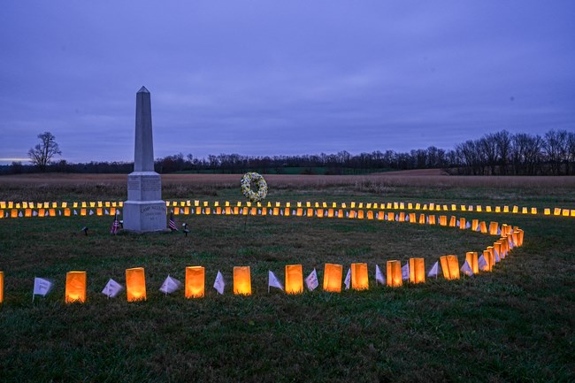 A stone obelisk in a grass cemetery surrounded by light lumianria bags.