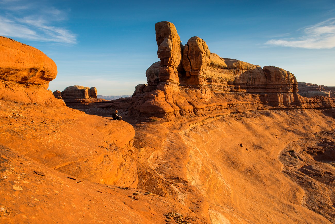 A person sits on a saddle between two canyons. The rock is orange and brown with a blue sky above.