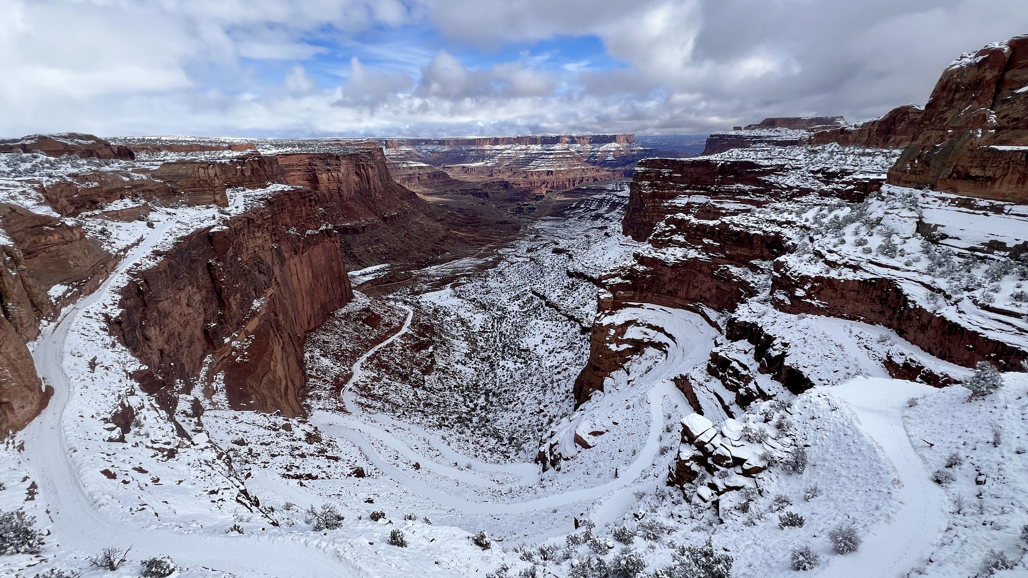 Snowy scene of a dirt road with switchbacks curving downward towards the bottom of a reddish-brown canyon. Cloudy sky above.