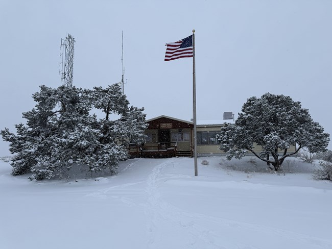 Snowy scene in front of a wooden trailer ranger station. Trees stand on either side of the building. Snow covers the ground and a flag pole rises into a grey sky.