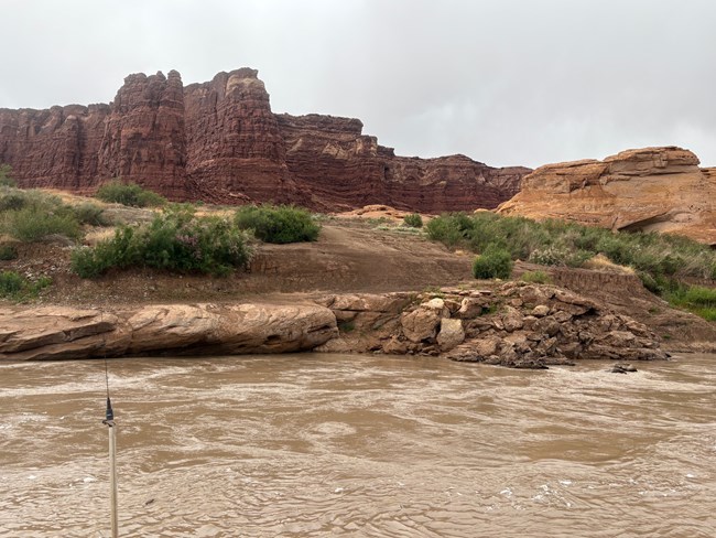 View from a boat showing river with brown water. A rocky bank leads up to a dirt ramp. Green vegetation grows on the bank. Red cliffs are in the background.
