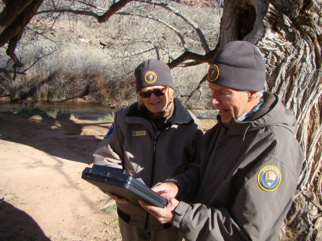 Two people standing outside in brown jackets and hats with volunteer patches holding a clipboard.