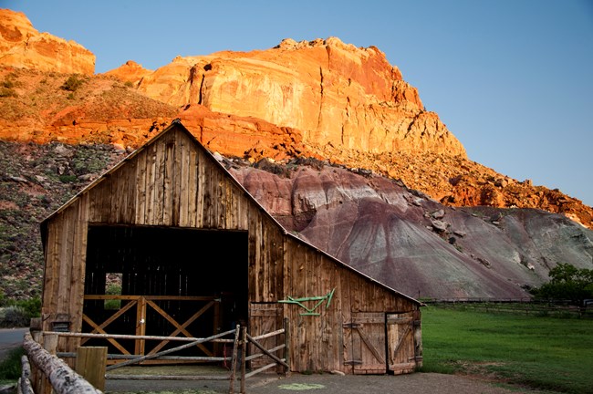 Large wooden barn with green pasture to the right of it and colorful red and gray striped cliffs above.