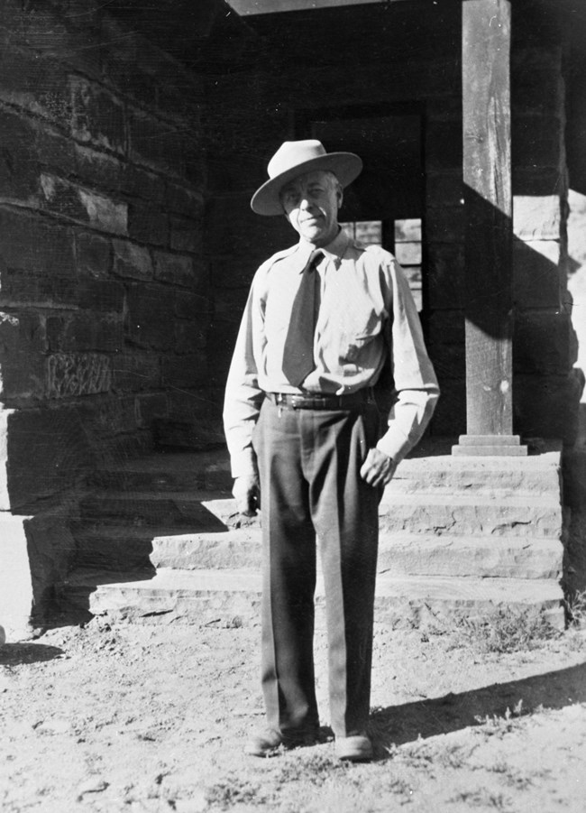 Black and white photo of man wearing a park service uniform and flat hat, standing in front of a stone building.