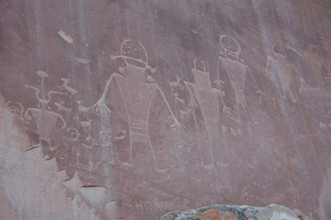 Five human-like figures pecked into stone, with smaller images, including bighorn sheep, interspersed between them, on reddish brown rock.