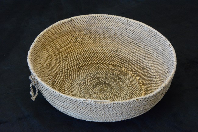 Round, woven, tan colored basket on a black background.