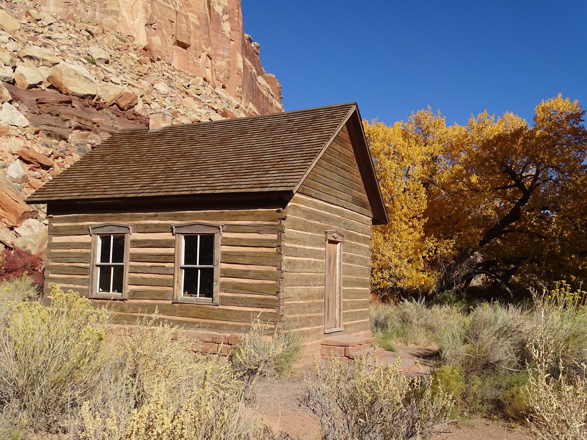 Log cabin building with stone foundation, in front of red cliffs, yellow trees, and blue sky.