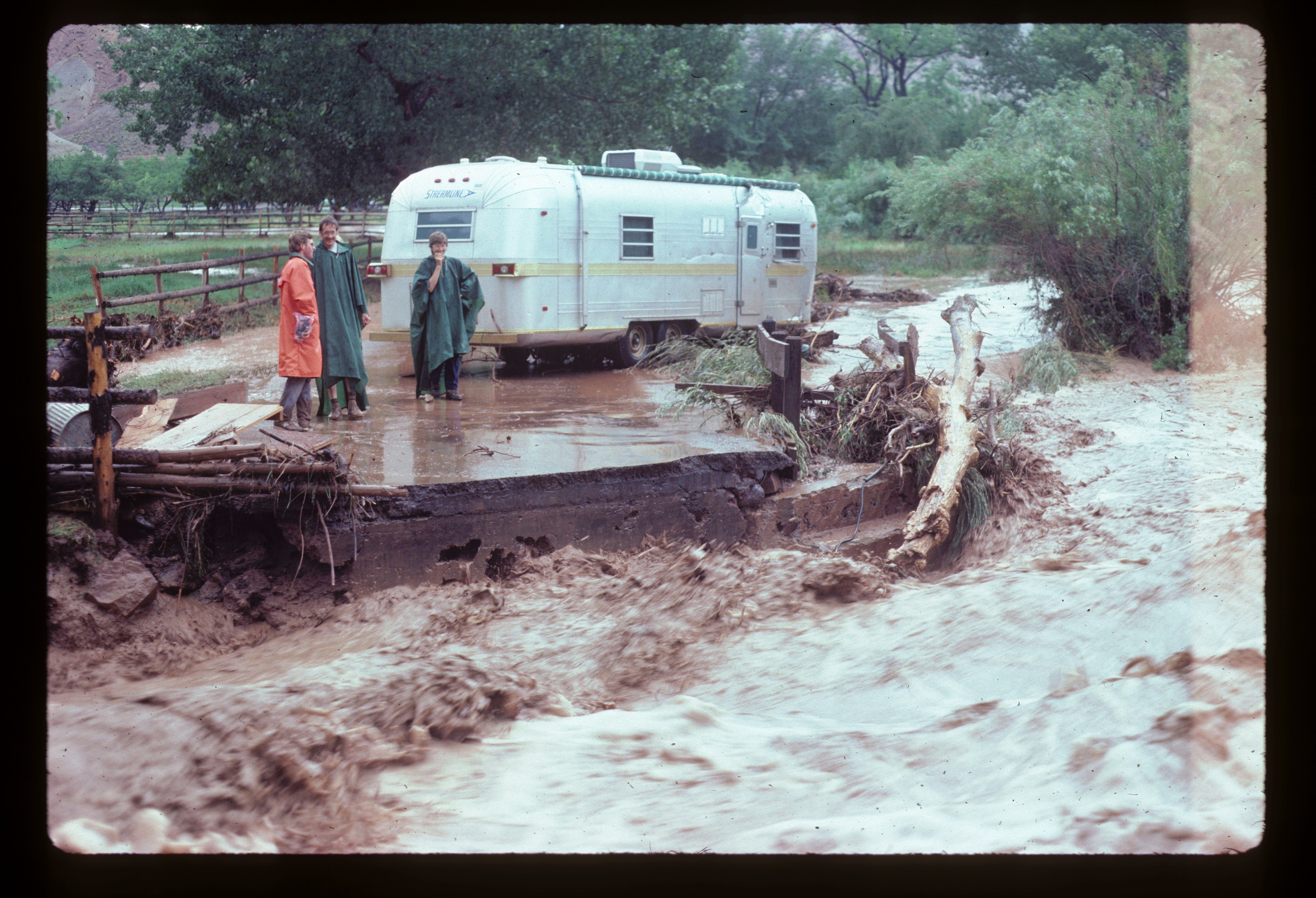 Visitors in ponchos in front of an RV as a flash flood roars across a collapsed road in front of them