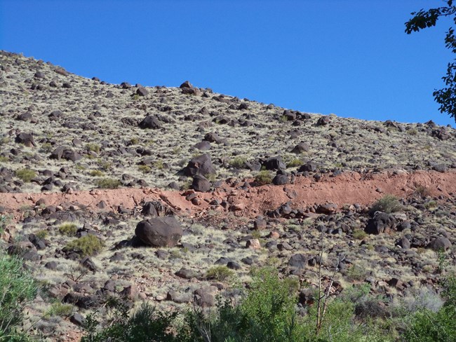 Large black boulders scattered on a grassy green hillside, with a large disturbed area crossing the mesa, where upturned dirt is visible. Blue skies above.