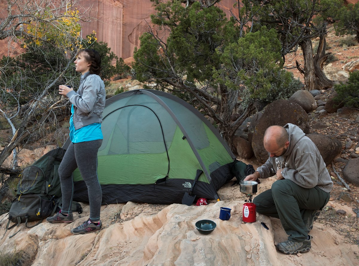 Two people, a tent, camp stove and equipment on tan sandstone in a red rock canyon with some trees.