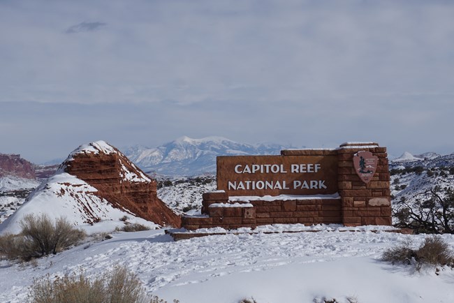 Snow-covered large stone sign with NPS arrowhead and "Capitol Reef National Park" on it, with a backdrop of snowcovered red slopes and snowy mountains, with a cloudy sky.