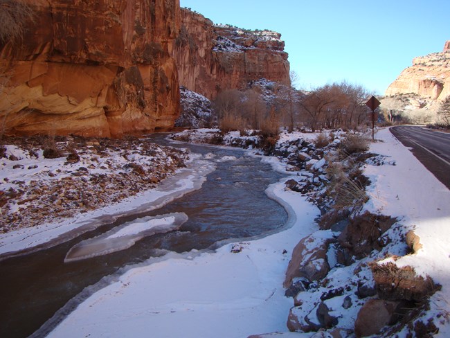 Small river with ice along the edges and in the center, and snow on the riverbanks, below red cliffs and blue sky, with a road to the right of the river.