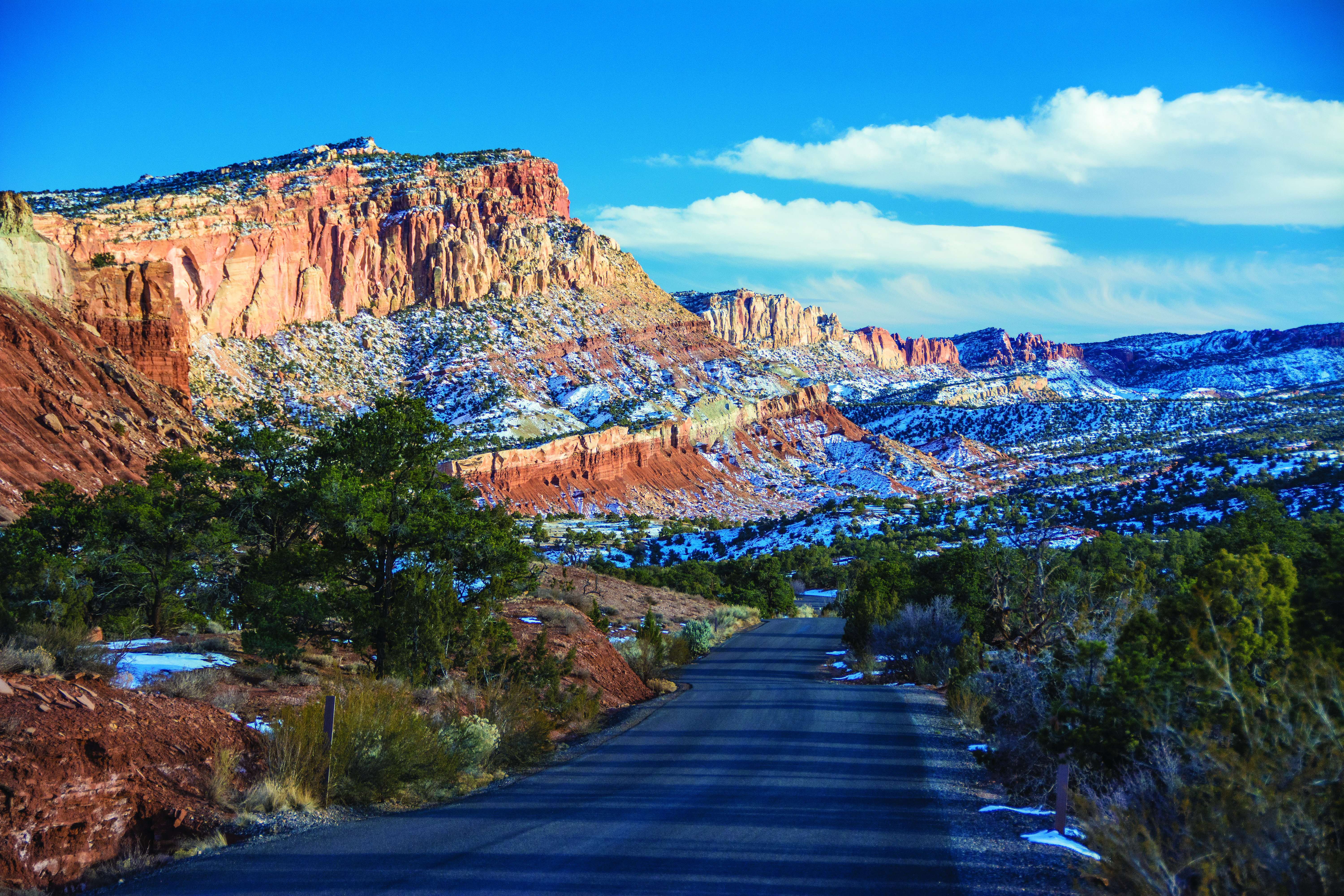 Blacktop road with small green trees and shrubs and red cliffs covered in snow, with blue sky and clouds above.