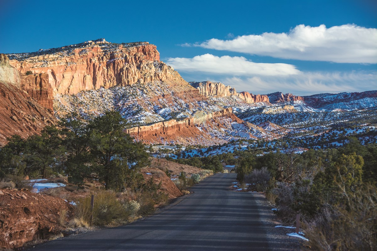 Blacktop road with small green trees and shrubs and red cliffs covered in snow, with blue sky and clouds above.