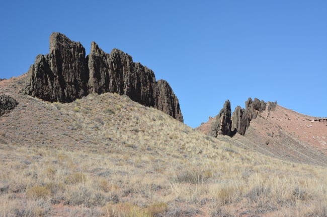 long line of jagged black rocks rising out of eroding red hills with blue sky above.