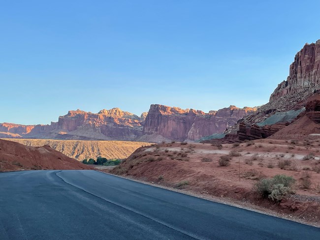 View from atop a hill on a road with a line of red sandstone cliffs in sunlight in the distance.