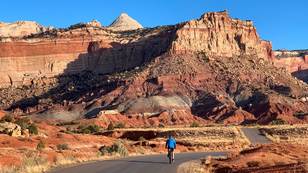 Cyclist riding along the scenic drive road through Capitol Reef National Park, with colorful layered cliffs in the distance under a clear blue sky