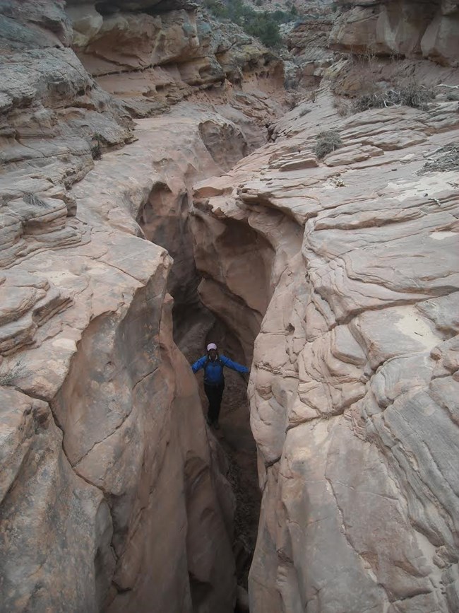 Person in the bottom of a slot canyon, looking up at the camera, with a wider, taller canyon above.