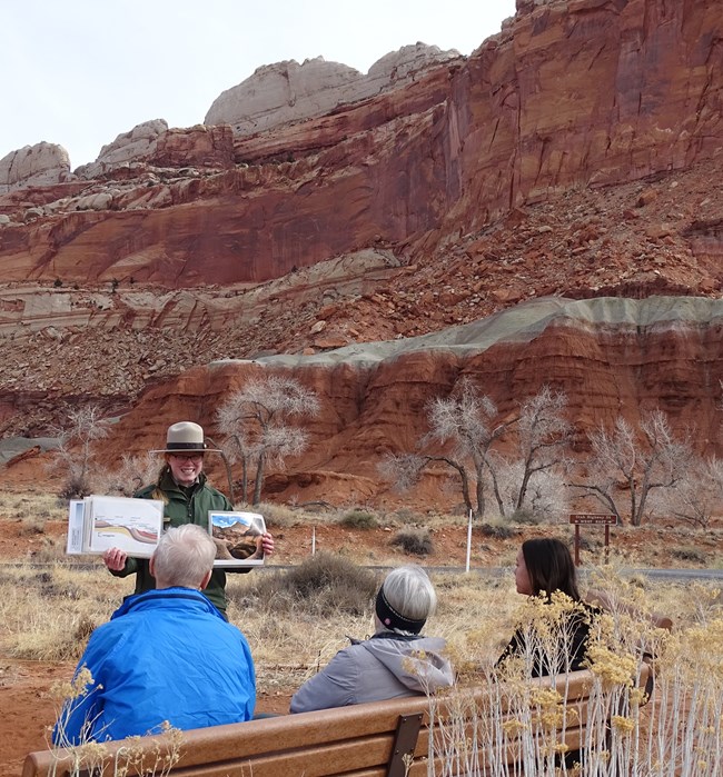 Ranger in flat hat, holding photos and diagrams up in front of audience of three people, with a background of colorful rock layers and formations.