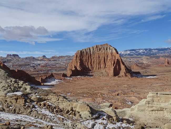 Large orange monolith with colorful rock cliffs surrounding the valley it is in, with blue sky, clouds, and a bit of snow.
