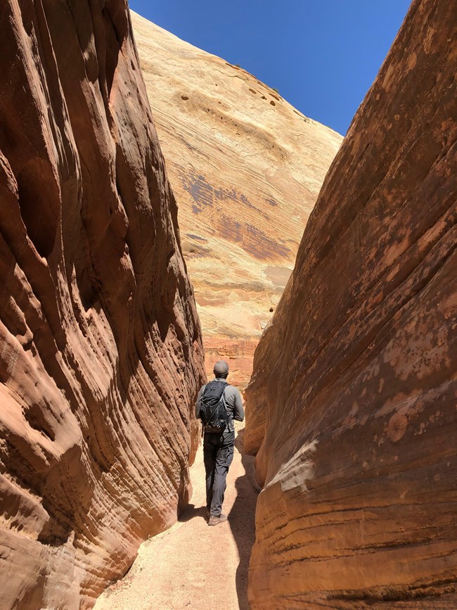 Person wearing a backpack stands in slot canyon, with red-orange cliffs on both sides, and blue sky above.