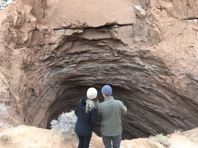 Two people looking into a large, dark hole at the base of a cliff.