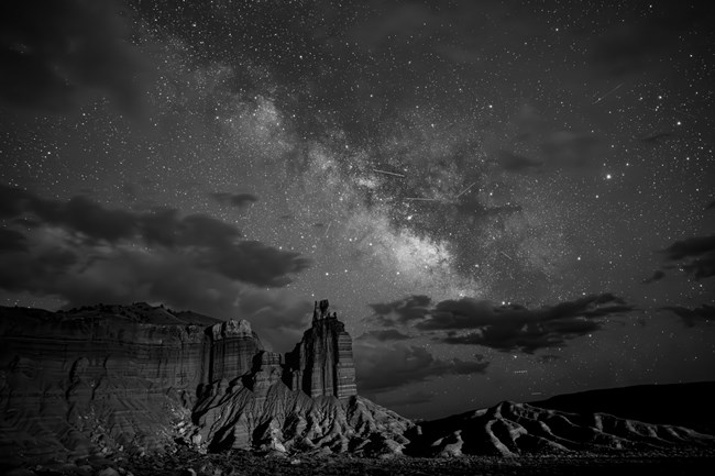 Black and Gray photo of the Milky Way over Chimney Rock. Several streaks of light from meteors fly across the sky.