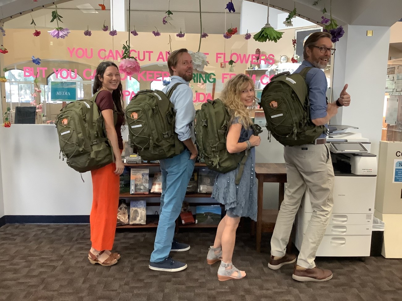 Four adults stand in the library carrying green NPS backpacks.