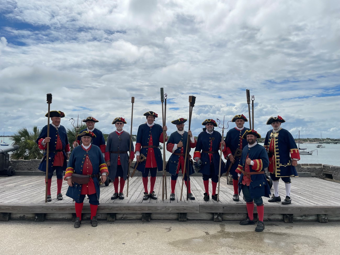 10 people dressed in blue and red colonial Spanish uniforms. 7 of them hold long-handled tools for loading and cleaning the cannons.