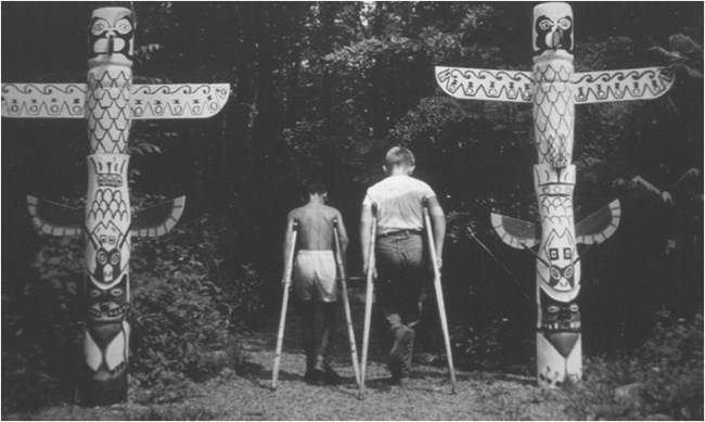 Two boys on crutches walk between 2 totem poles