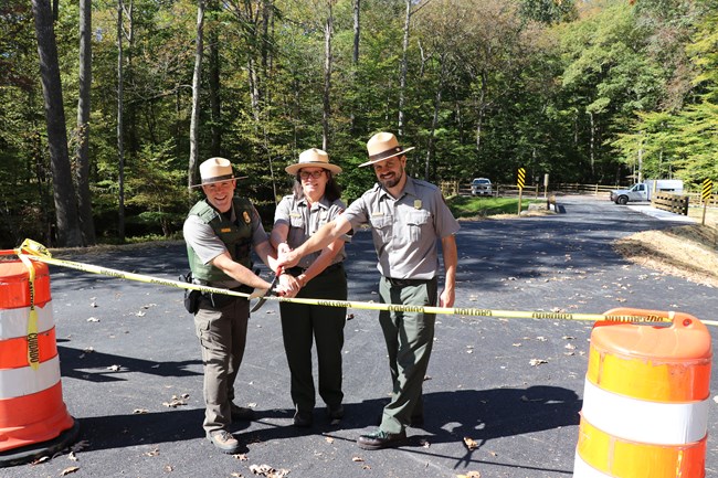 three park rangers wearing uniforms and flat hats hold an oversized pair of scissors to cut caution tape. The rangers are two men and a woman, and behind them is a newly constructed bridge.