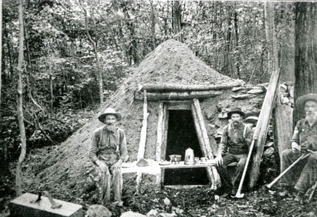 Historical photograph of three colliers sitting next to their hut made of sticks.