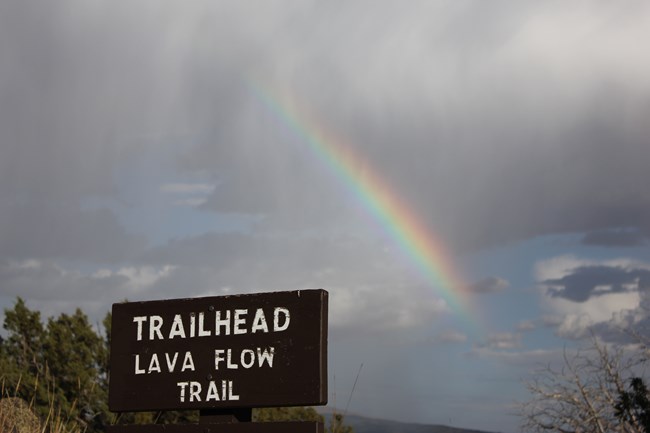 A wooden trail sign with words etched as "Trailhead.  Lava Flow Trail".  Trees are in the nearby horizon below a dark cloudy sky where a partial rainbow cascades down to the right.
